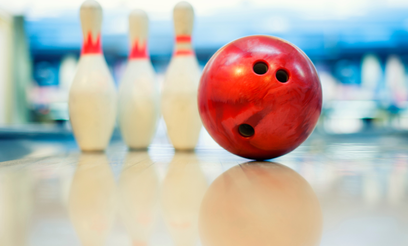 A shiny red bowling ball and three white bowling pins sitting on a brightly-lit bowling lane.