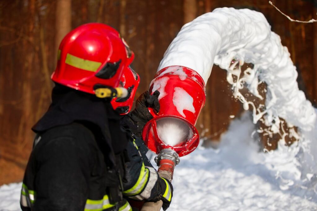 Firefighter in red hat holding red fire hose with firefighting foam coming out.