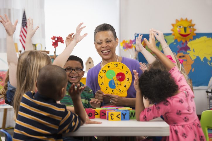 Child care provider holding clock surrounded by children raising their hands