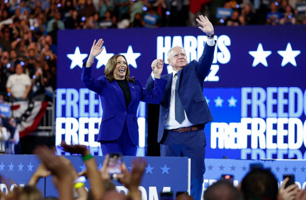 Kamala Harris and Tim Walz waving to crowd in front of blue sign that reads "Freedom" at Milwaukee event.