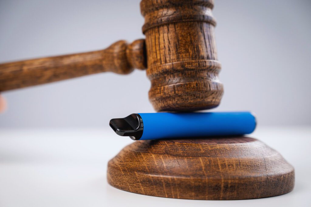 Judge hammering disposable vape with referee's gavel on white table.