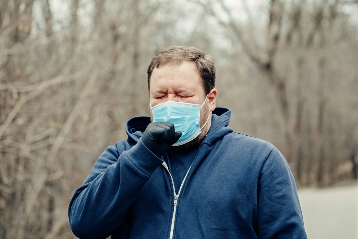 Man wearing a blue hooded sweatshirt, black gloves, and a mask, coughing into his hand.
