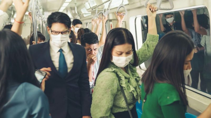 People wearing masks on a train, with a man unmasked and coughing in the background.