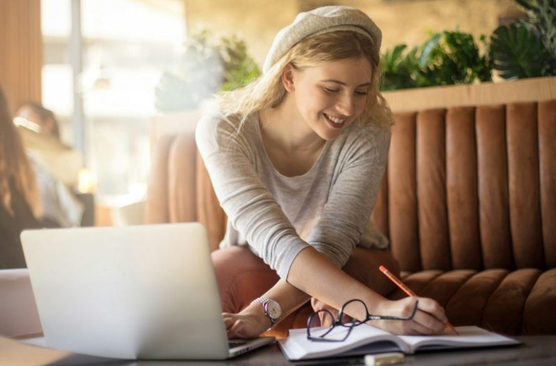 Blond woman smiling and writing something in a notebook while sitting in front of a laptop.