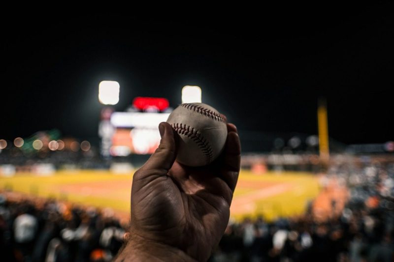 a hand holds a baseball with a baseball field in the background