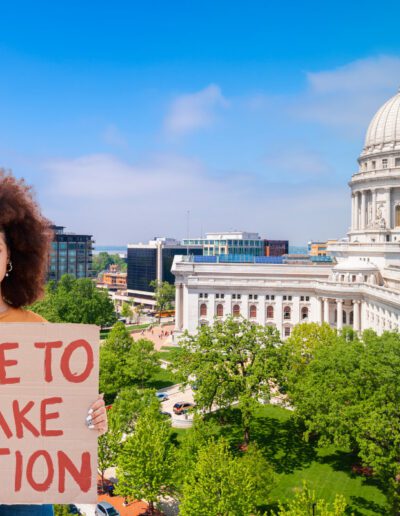 Photo illustration. Woman holding protests sign reading "time to take action". Madison Wisconsin in background